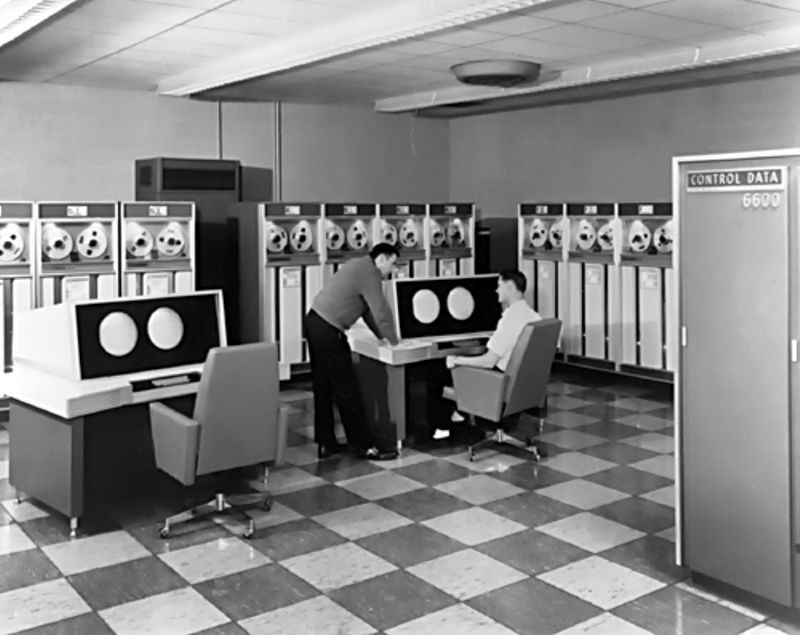 Two men operating the dual-console of the CDC 6600, with tape drives in the background.